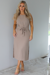 Woman wearing a tan, high neckline, midi dress that has a tie detail at the waistline and split hem details on either side. The photo is taken indoors against a white wall with white marble flooring. 