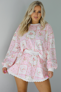 Woman wearing a two piece set featuring a pink floral set that has an oversized long sleeved top and high waisted skirt. The photo is taken indoors against a white wall.