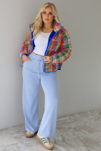 Woman wearing a multi-colored plaid, quilted style jacket paired with a white top underneath and high waisted blue lounge pants. The photo is taken indoors against a white wall with white marble flooring. 