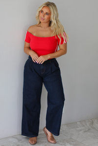 A blonde woman modeling a bright red off-the-shoulder short-sleeve top with white stripes on the sleeves, paired with dark wide-leg denim jeans, standing indoors against a white wall with white marble flooring. 