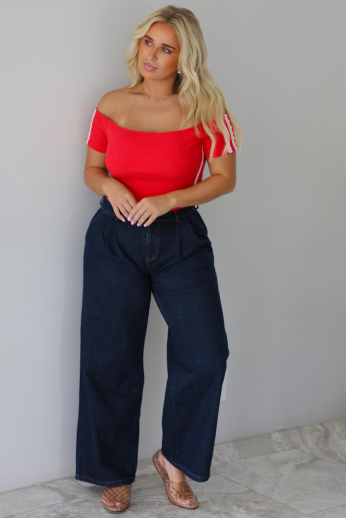 A blonde woman modeling a bright red off-the-shoulder short-sleeve top with white stripes on the sleeves, paired with dark wide-leg denim jeans, standing indoors against a white wall with white marble flooring. 