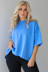 Woman wearing an oversized blue box tee with black leggings. The photo shows a closeup of the tee. The photo is taken indoors against a white wall with white marble flooring. 