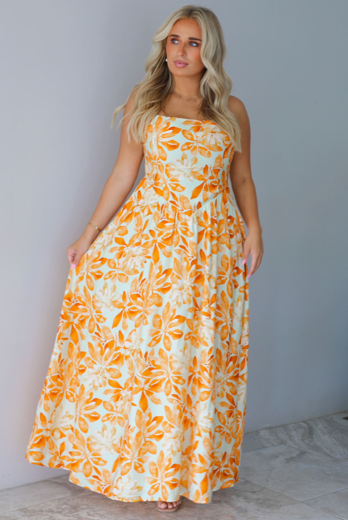 A woman modeling a spaghetti strapped, floor-length maxi dress with a white base and a vibrant marigold orange floral pattern, standing indoors against a white wall with white marble flooring. 