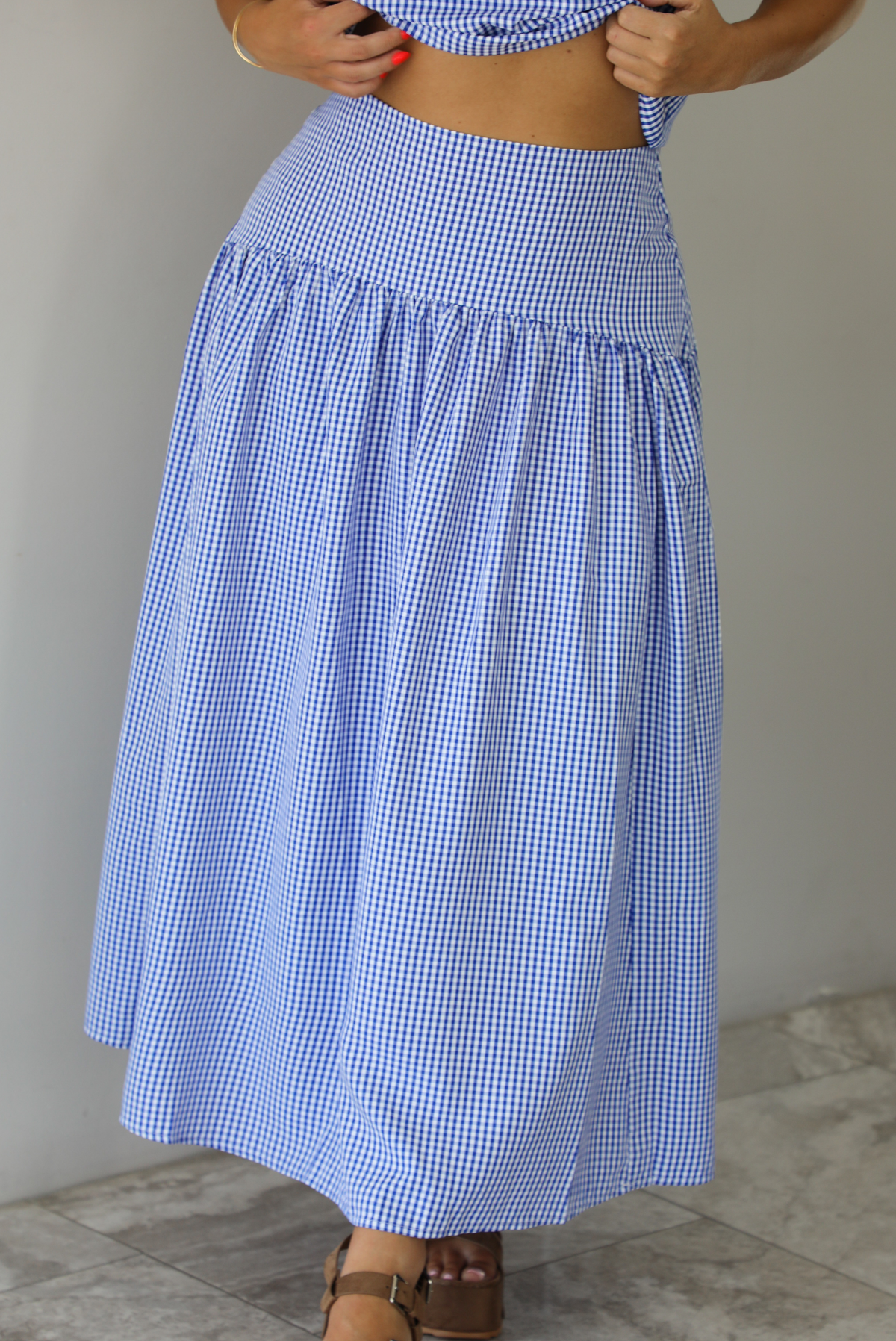 Woman wearing a blue and white gingham two piece set featuring a sleeveless top and high waisted, maxi length skirt indoors against a white wall with white marble flooring. The photo shows a closeup of the front of the skirt. 