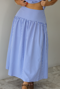 Woman wearing a blue and white gingham two piece set featuring a sleeveless top and high waisted, maxi length skirt indoors against a white wall with white marble flooring. The photo shows a closeup of the front of the skirt. 