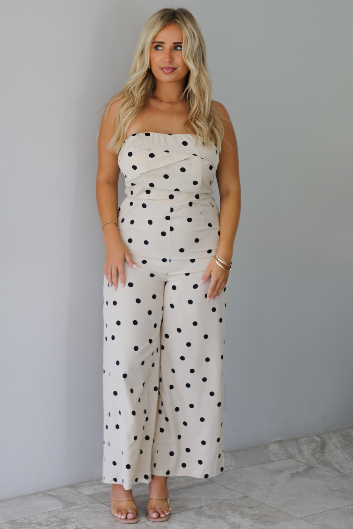 A woman wearing a sand strapless jumpsuit with a black polka dot print pattern throughout, standing indoors against a white wall with white marble flooring. 
