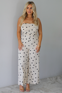 A woman wearing a sand strapless jumpsuit with a black polka dot print pattern throughout, standing indoors against a white wall with white marble flooring. 
