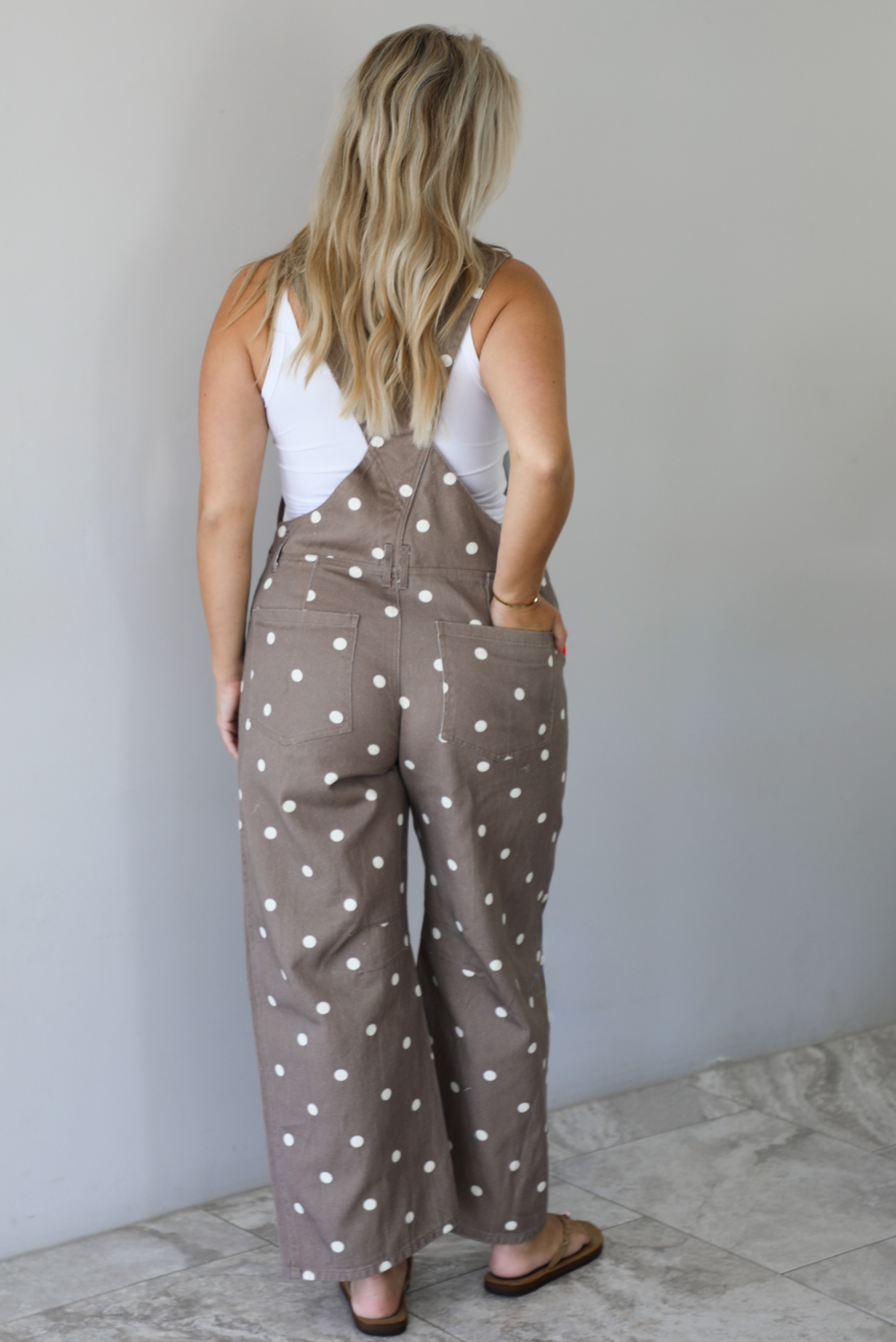 Woman wearing a brown with white polka dots overalls jumpsuit indoors against a white wall with white marble flooring. The photo shows the back of the overalls.