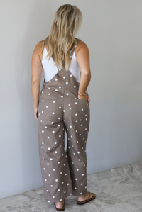 Woman wearing a brown with white polka dots overalls jumpsuit indoors against a white wall with white marble flooring. The photo shows the back of the overalls.