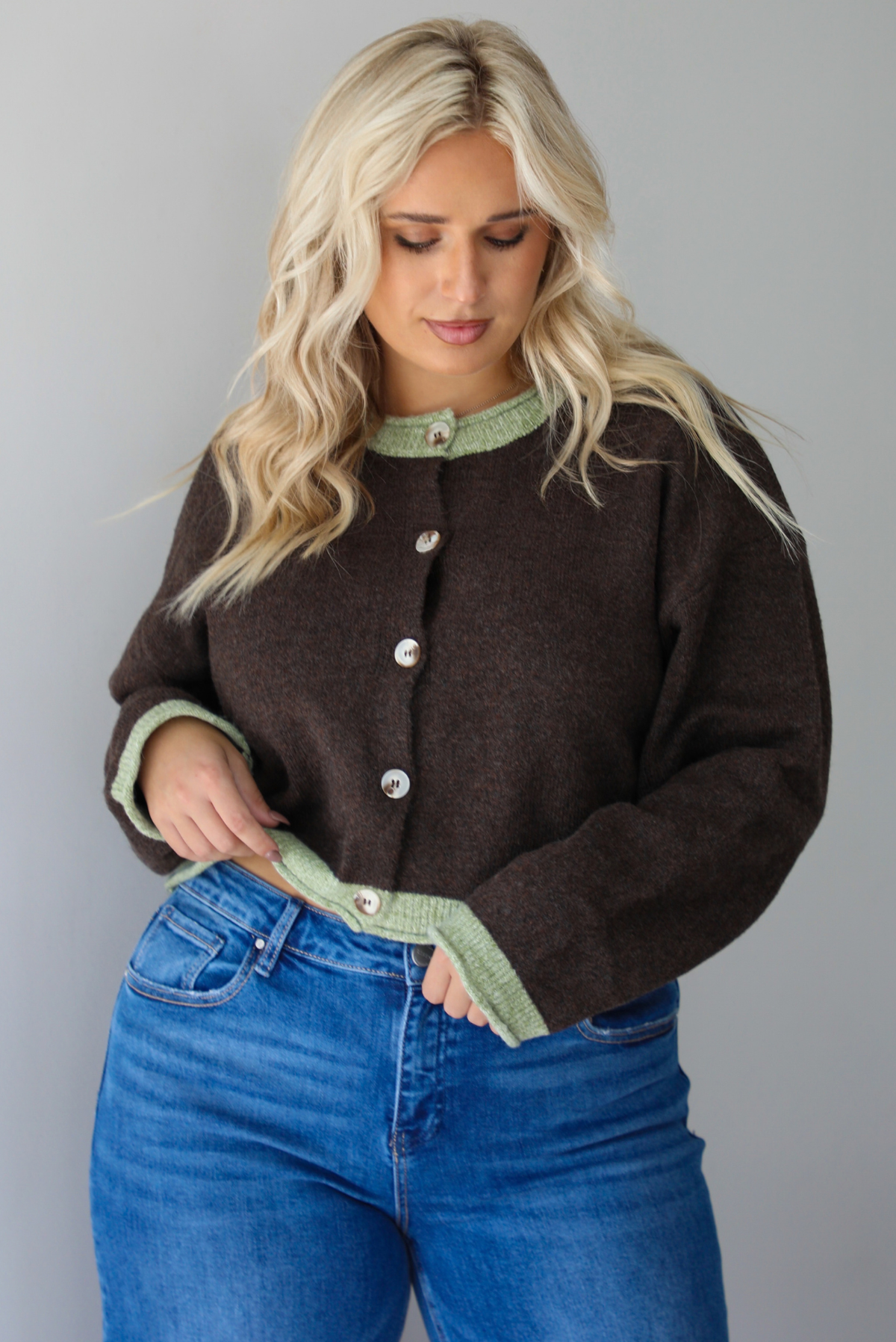 A woman wearing a dark brown with green trim cardigan fully buttoned up paired with dark wash denim jeans, standing indoors against a white wall.