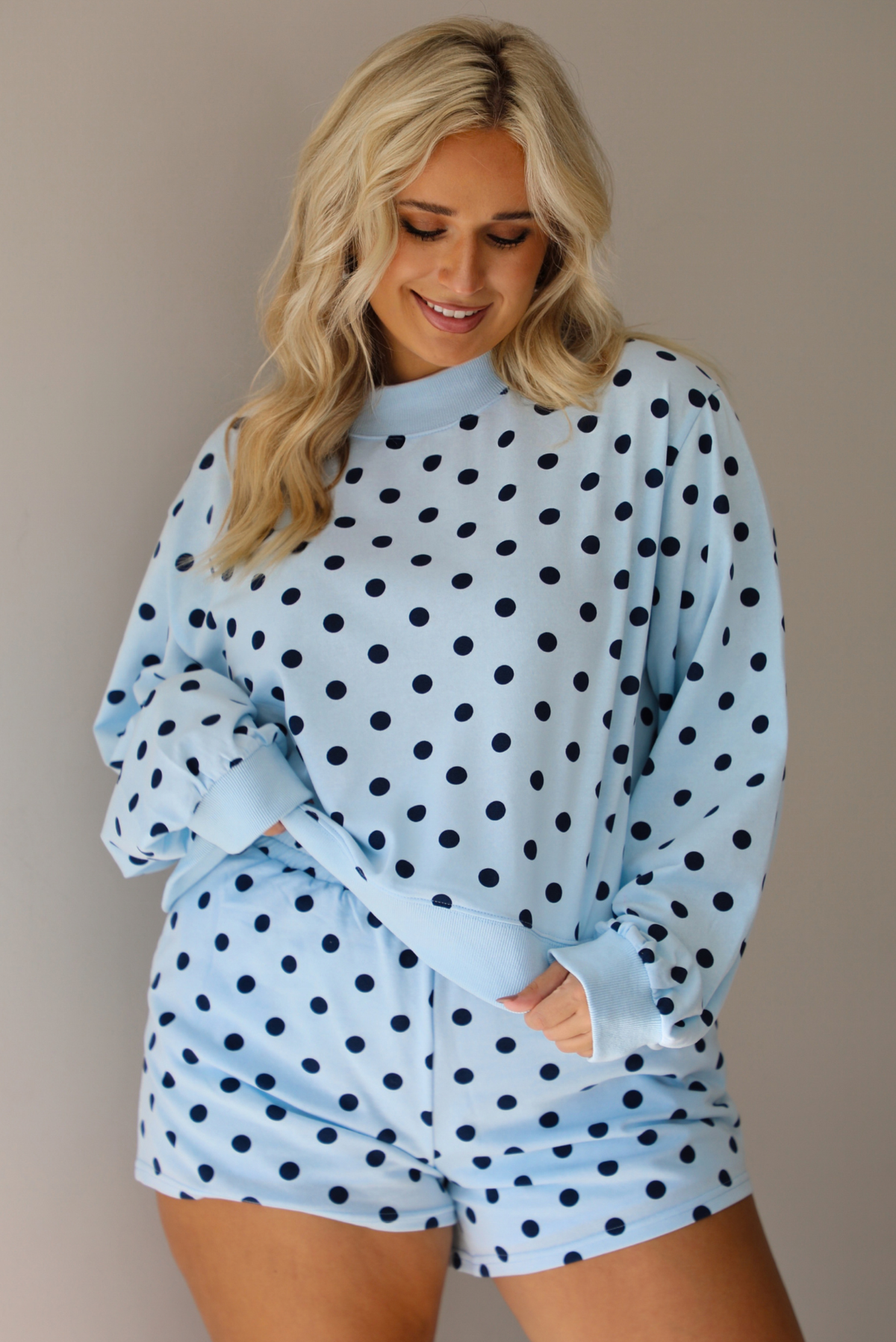 A woman wearing a blue two piece set with a navy polka dot pattern throughout, featuring a sweatshirt and high waisted sweat-shorts, shown indoors against a white wall.