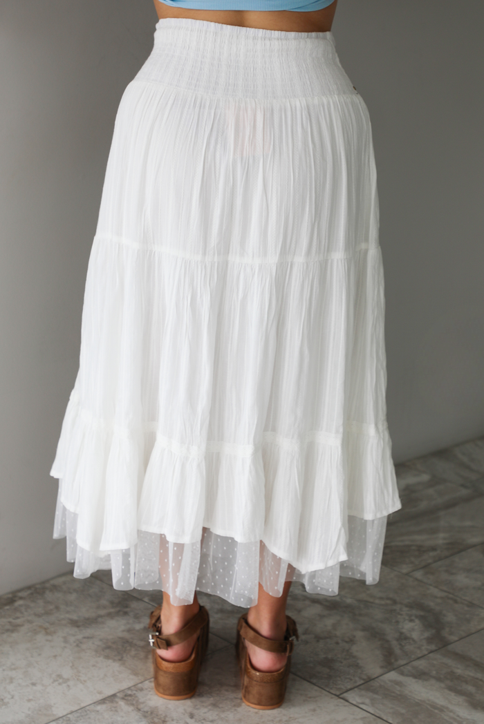 Woman wearing a white tiered mai length skirt with studded details at the waistline and sheer mesh detailing at the hem indoors against a white wall with white marble flooring. The photo shows the back of the skirt.