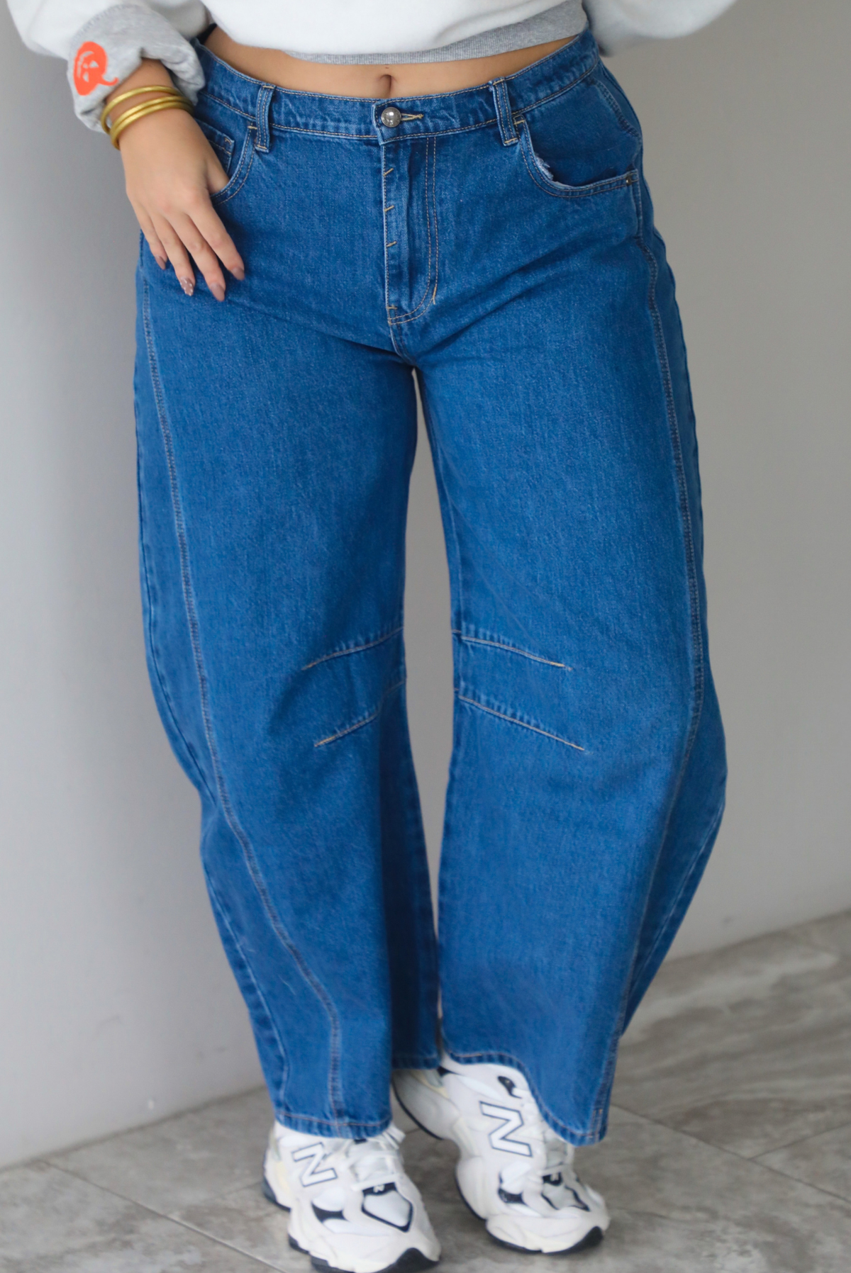 A woman shown from the waist down, focusing on dark wash barrel denim jeans that have a mid-rise waistline, paired with a grey sweatshirt and New Balance chunky sneakers, standing indoors against a white wall with white marble flooring.  