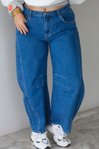 A woman shown from the waist down, focusing on dark wash barrel denim jeans that have a mid-rise waistline, paired with a grey sweatshirt and New Balance chunky sneakers, standing indoors against a white wall with white marble flooring.  