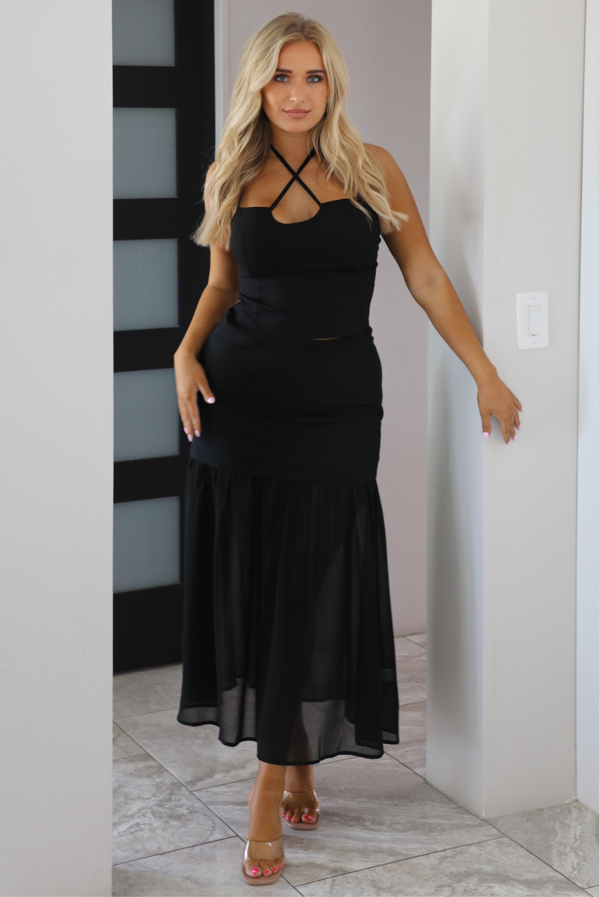 A woman modeling a black set with a strappy halter-style top and a flowing skirt, standing indoors against a light wall.