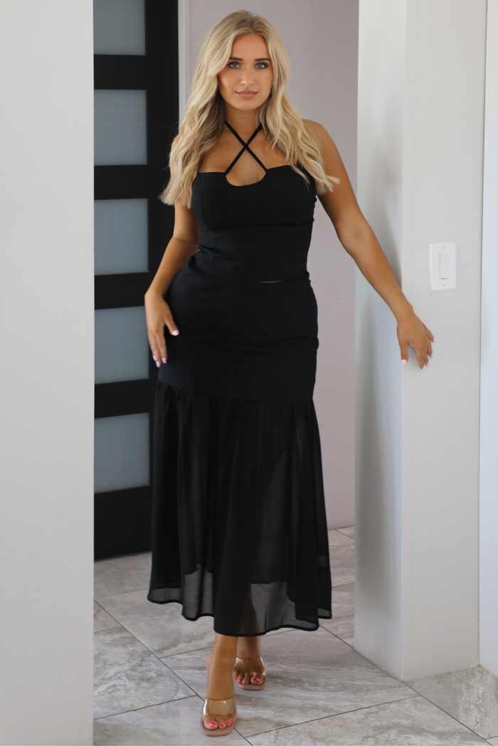 A woman modeling a black set with a strappy halter-style top and a flowing skirt, standing indoors against a light wall.