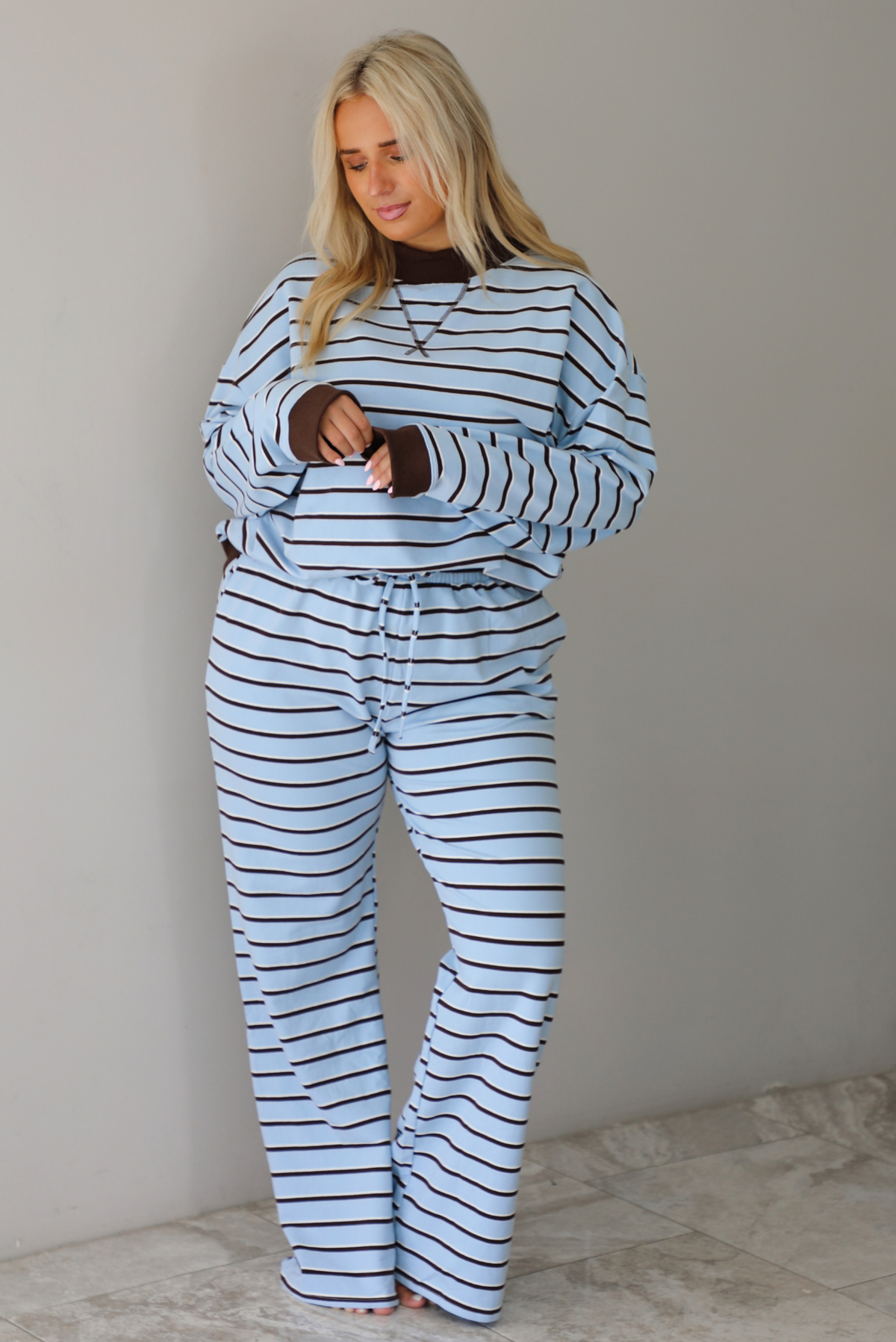 A woman wearing a blue matching two piece set with dark brown and white striped details, featuring an oversized long sleeved top and high waisted loose fitting, flared leg pants, standing indoors in front of a white wall with white marble flooring. 