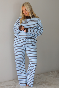 A woman wearing a blue matching two piece set with dark brown and white striped details, featuring an oversized long sleeved top and high waisted loose fitting, flared leg pants, standing indoors in front of a white wall with white marble flooring. 