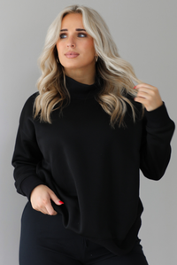Woman wearing a black high-neck, oversized sweater and high waisted black pants. The photo is taken indoors against a white wall with white marbled flooring. 