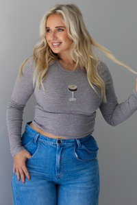 A woman wearing a striped, cropped long sleeved top with a beaded espresso martini on the front with medium wash denim jeans, standing indoors against a white wall.