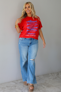 A woman wearing a red graphic t-shirt with blue sardine illustrations and the text 'Sardines LISBON FISH MARKET', paired with blue jeans, standing indoors against a white wall with white marble flooring.