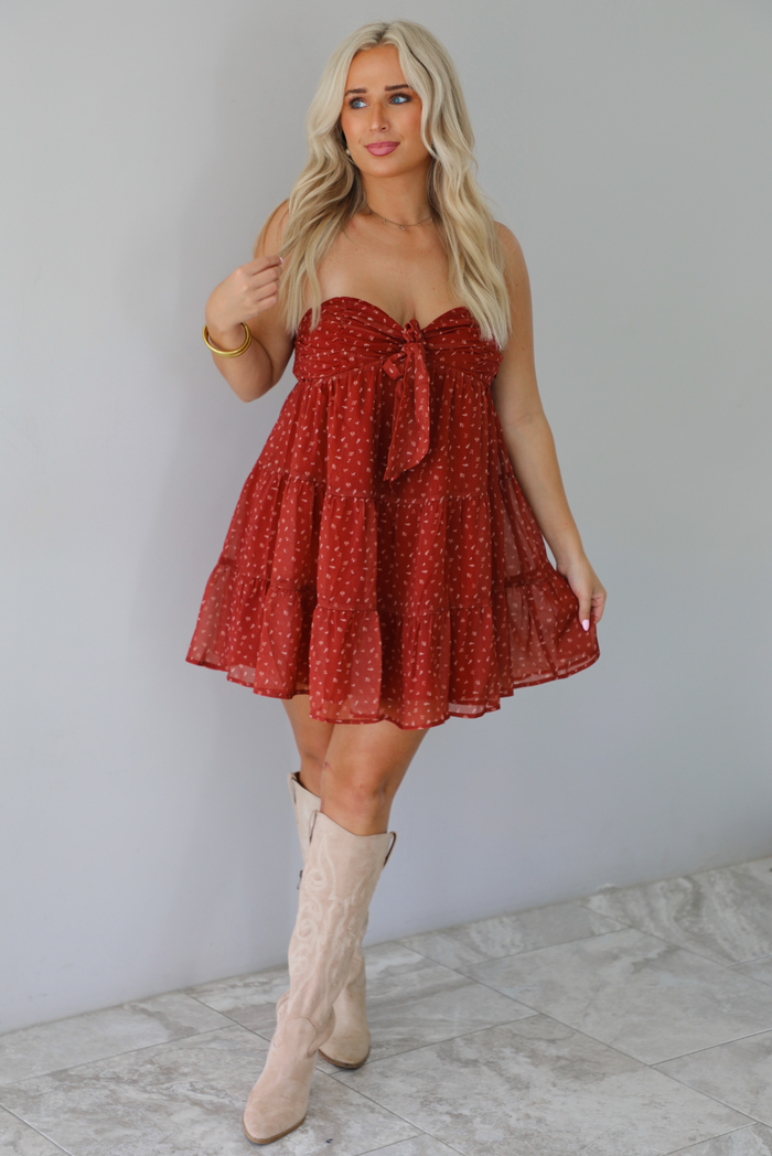 A woman wearing a rust mini length dress with a cream floral and leaf tiny print design throughout, has a strapless design with a tie detail in the center of the bust, with a baby-doll dress cut that's fitted at the bust with a flowy skirt, standing indoors against a white wall with white marble flooring. 