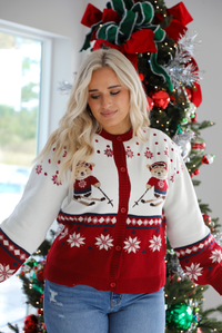 Woman model wearing a red and white Christmas cardigan with teddy bears skiing with functional button closures in the front. The cardigan is styled with medium wash denim jeans that are cuffed at the hem. The photo is taken inside a modern home with a decorated Christmas tree in the back. 
