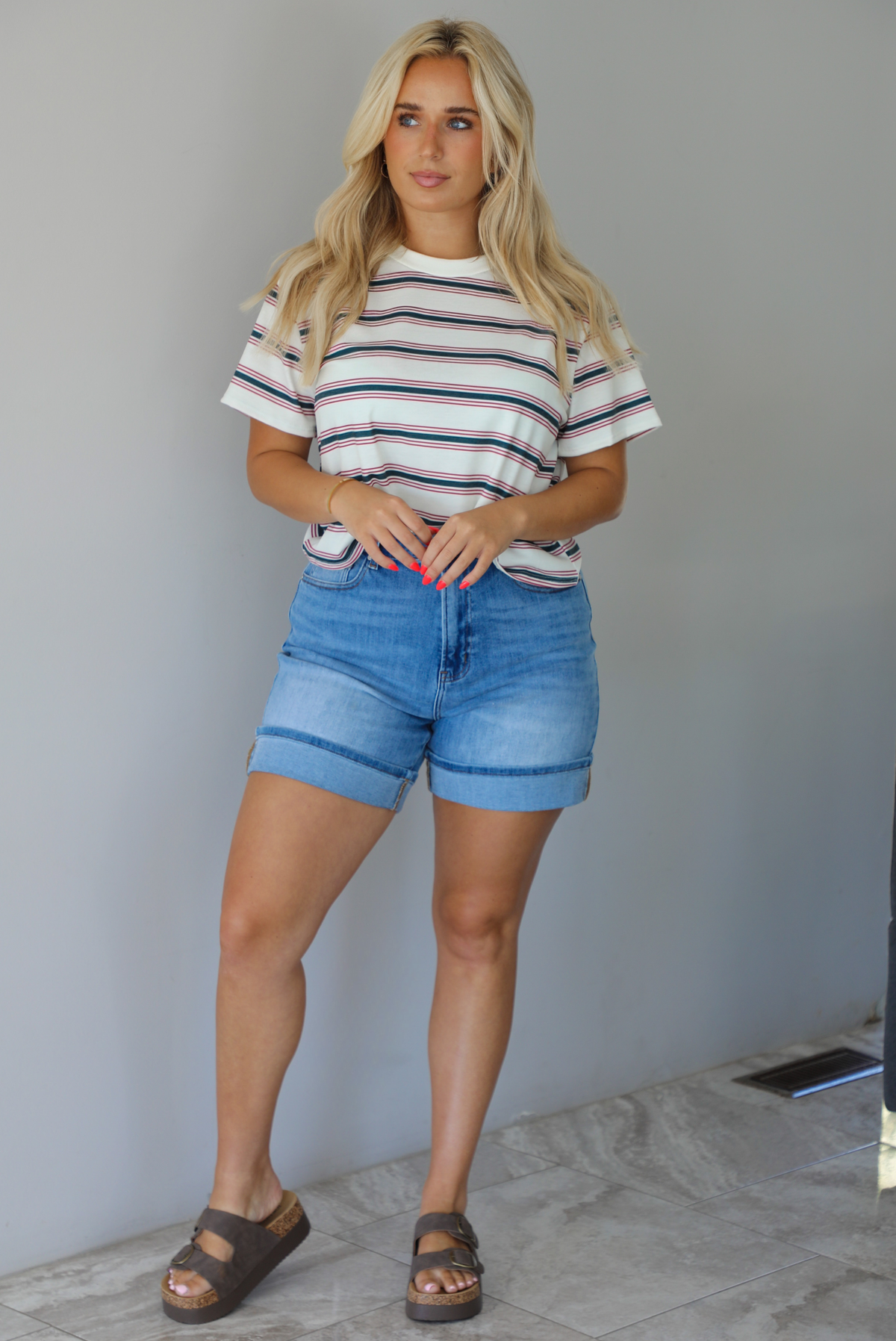 Woman wearing a striped, slightly cropped tee with light-wash denim shorts indoors against a white wall with white marble flooring. 
