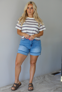 Woman wearing a striped, slightly cropped tee with light-wash denim shorts indoors against a white wall with white marble flooring. 