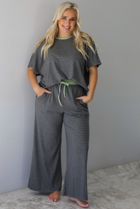 A woman wearing a charcoal and white striped two piece matching set with green trim detailing at the neckline and waistline, featuring an oversized short sleeved top and high waisted pants, standing indoors against a white wall with white marble flooring. 
