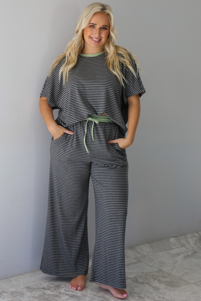 A woman wearing a charcoal and white striped two piece matching set with green trim detailing at the neckline and waistline, featuring an oversized short sleeved top and high waisted pants, standing indoors against a white wall with white marble flooring. 