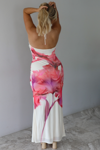 Woman wearing a cream maxi dress with a pink floral design, halter tie neckline, plunging neckline with a knotted detail at the upper waist indoors against a white wall with white marble flooring. The photo shows the back of the dress. 