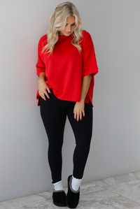 Woman wearing an oversized red t-shirt with black, fitted lettings, scrunchie socks, and black ugg boots. The photo is taken indoors against a white wall with white marbled flooring. 