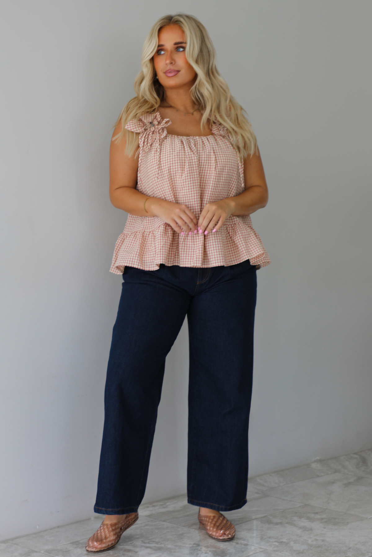 A woman wearing a light maroon and cream gingham flowy top with dark denim jeans, standing indoors against a white wall with white marble flooring.