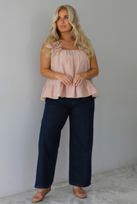 A woman wearing a light maroon and cream gingham flowy top with dark denim jeans, standing indoors against a white wall with white marble flooring.