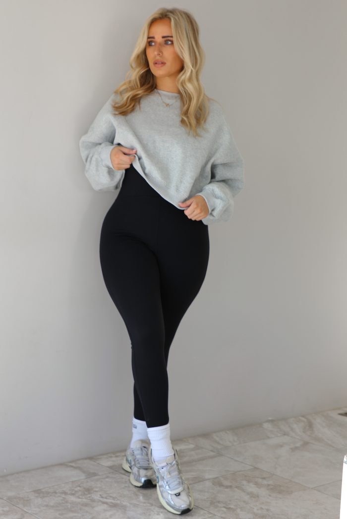 Woman wearing a heather grey cropped sweatshirt with a black fitted jumpsuit underneath. The outfit is paired with high ankle socks and silver sneakers. The photo is taken indoors against a white wall with white marble flooring. 
