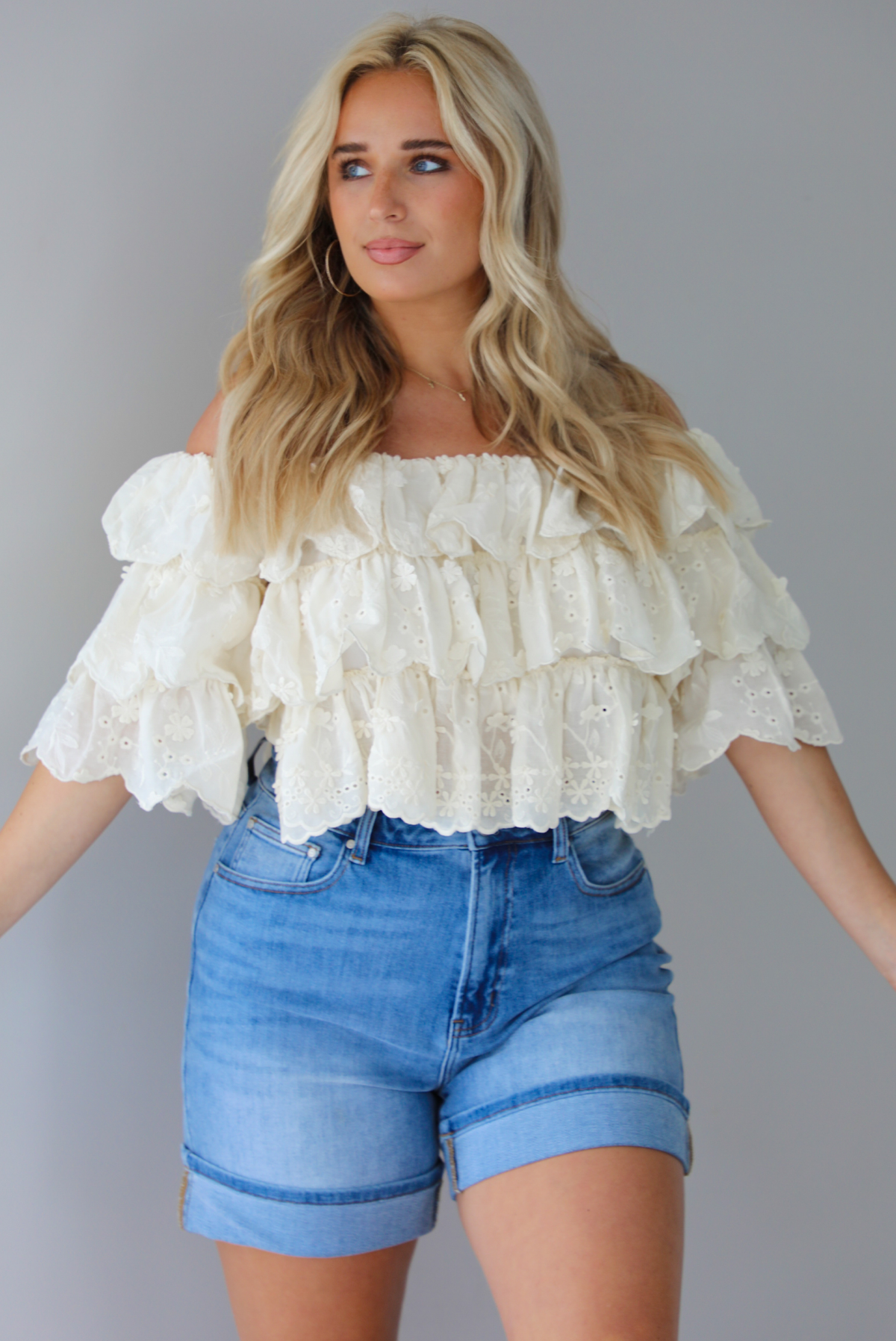 Woman wearing a cream off the shoulder, ruffled eyelet cropped top with medium wash denim shorts indoors against a white wall.