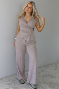 Woman wearing a taupe colored two piece set featuring a vest-appearance top with non-functional button closures on the left side and high waisted pants. The photo is taken indoors against a white wall with white marble flooring. 