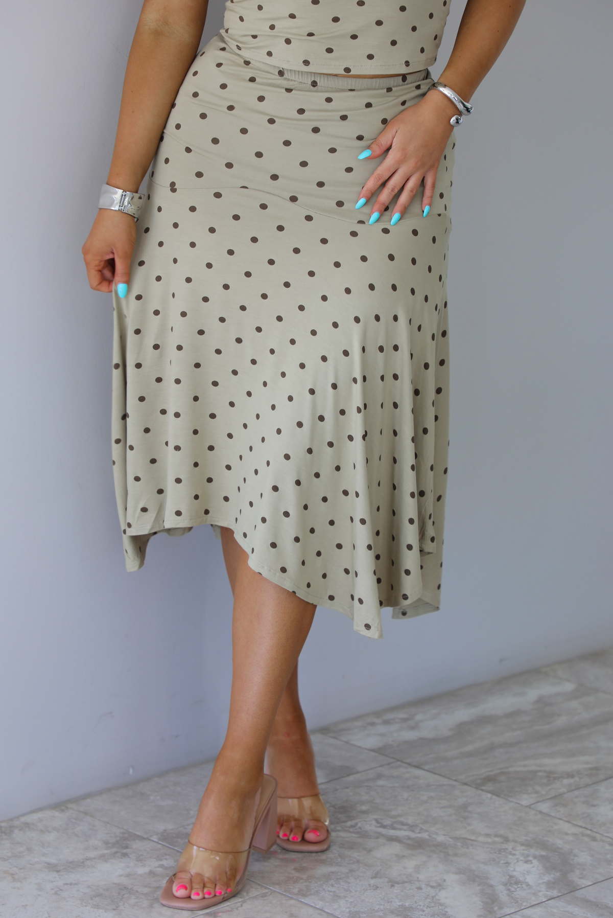Woman wearing a beige cropped tank and midi length skirt set with a brown polka dot print pattern. The photo is taken indoors against a white wall with white marble flooring and shows a closeup of the skirt, which has a high, elastic waistline, and an asymmetrical hemline. 