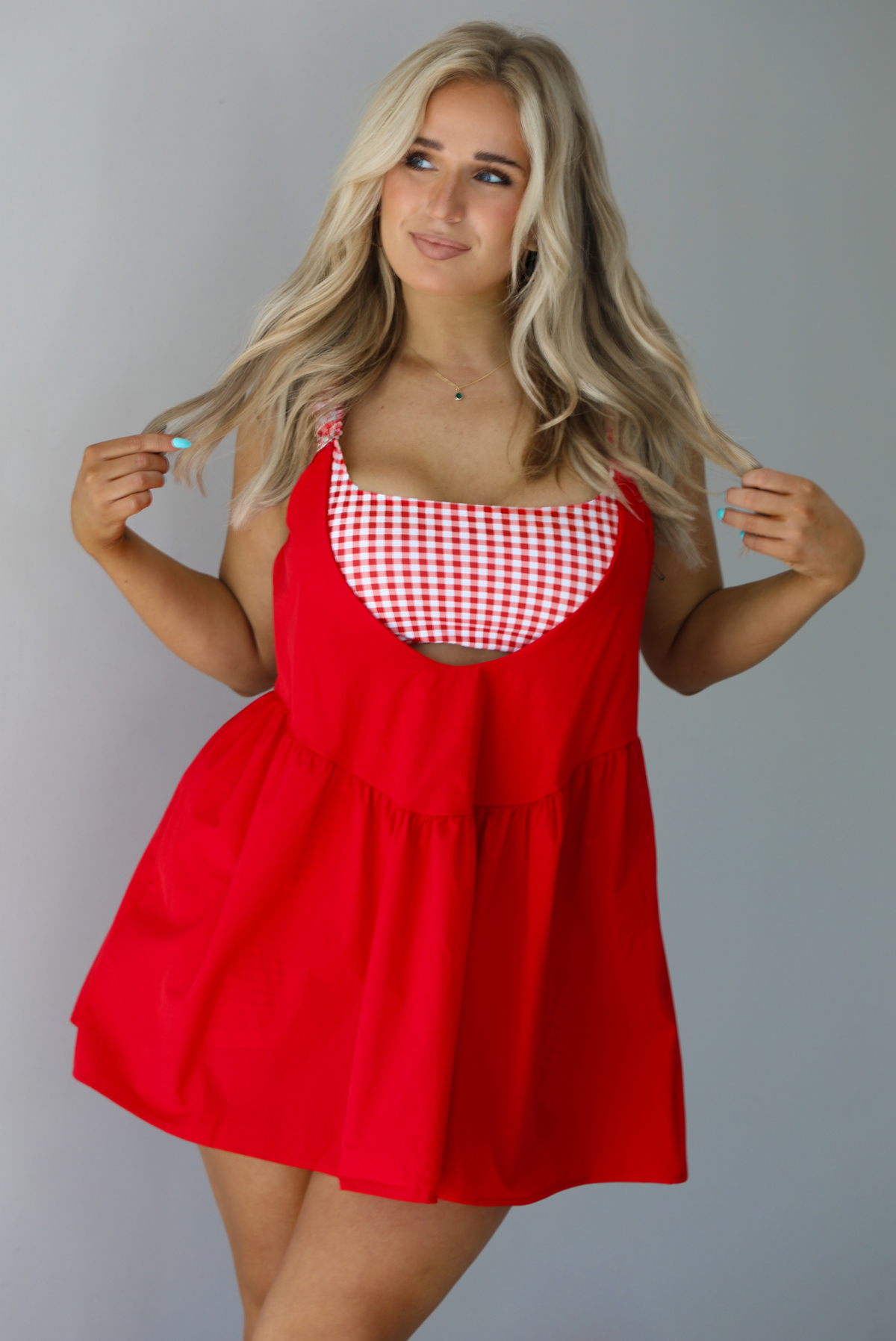 Woman wearing a red dress with a red and white gingham romper underneath. The photo is taken indoors against a white wall.
