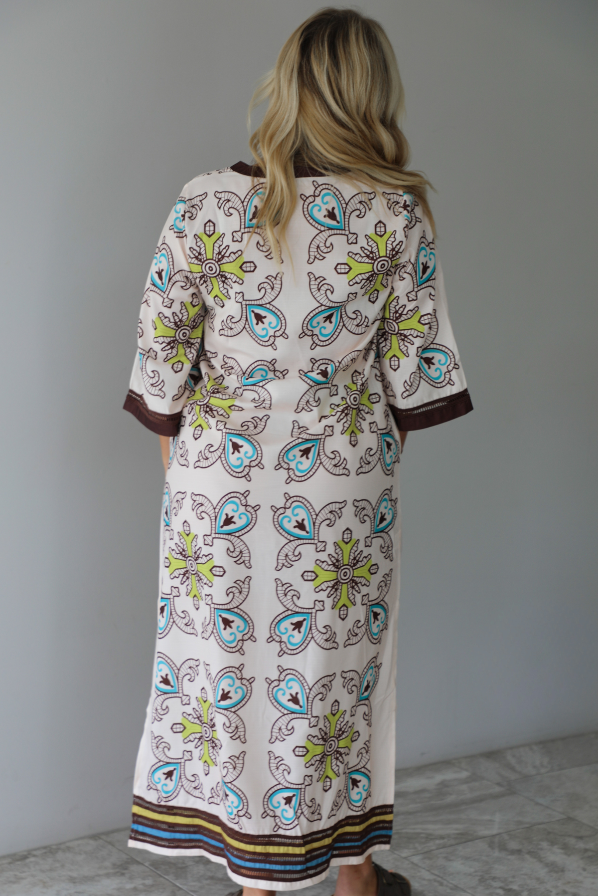 A woman shown from behind modeling a long kaftan dress with a colorful paisley pattern on a cream background, featuring brown trim and sandals, standing indoors against a white wall with white marble flooring. 
