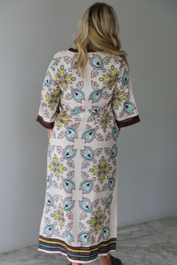 A woman shown from behind modeling a long kaftan dress with a colorful paisley pattern on a cream background, featuring brown trim and sandals, standing indoors against a white wall with white marble flooring. 