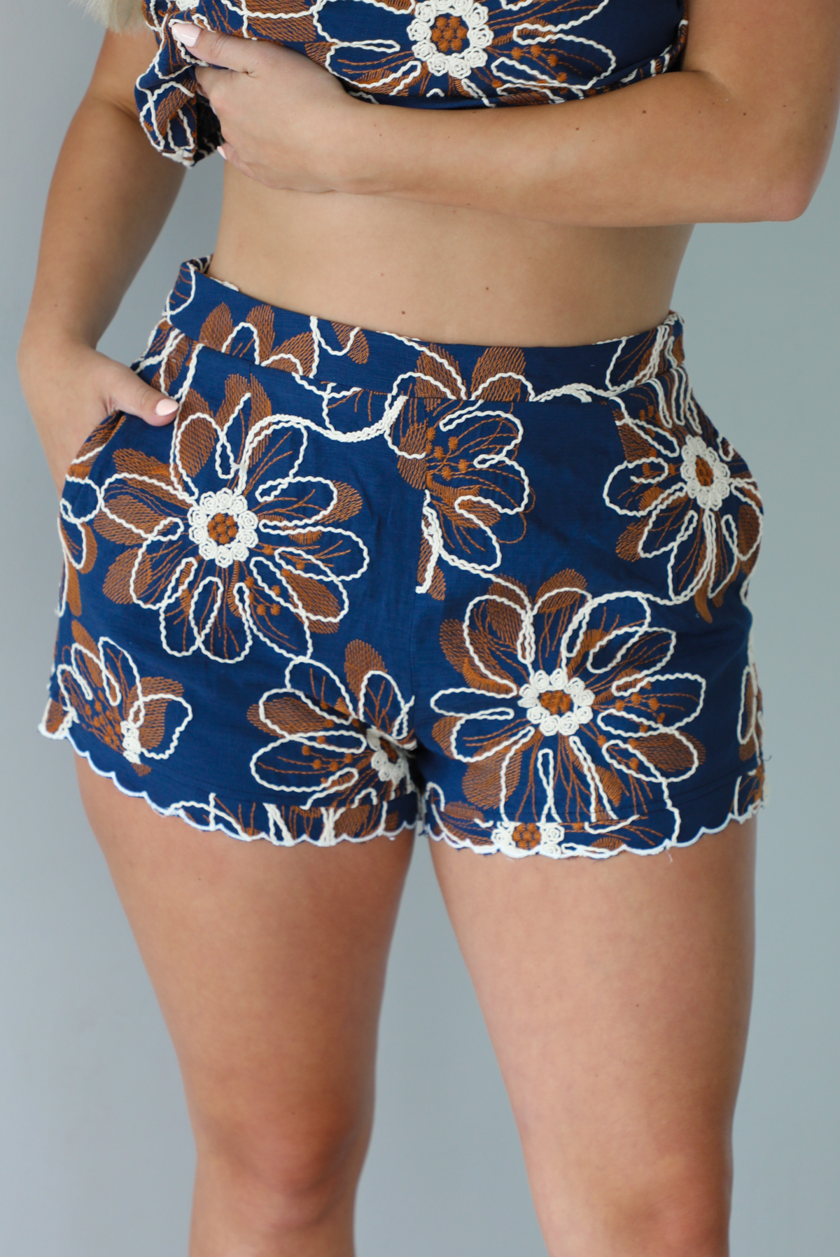 Woman wearing a navy set with a white and orange embroidered floral pattern throughout featuring a cropped top and high waisted shorts indoors against a white wall. The photo shows a closeup of the front of the shorts, which have functional of either side. 