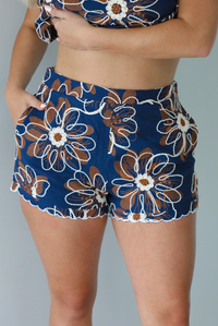 Woman wearing a navy set with a white and orange embroidered floral pattern throughout featuring a cropped top and high waisted shorts indoors against a white wall. The photo shows a closeup of the front of the shorts, which have functional of either side. 