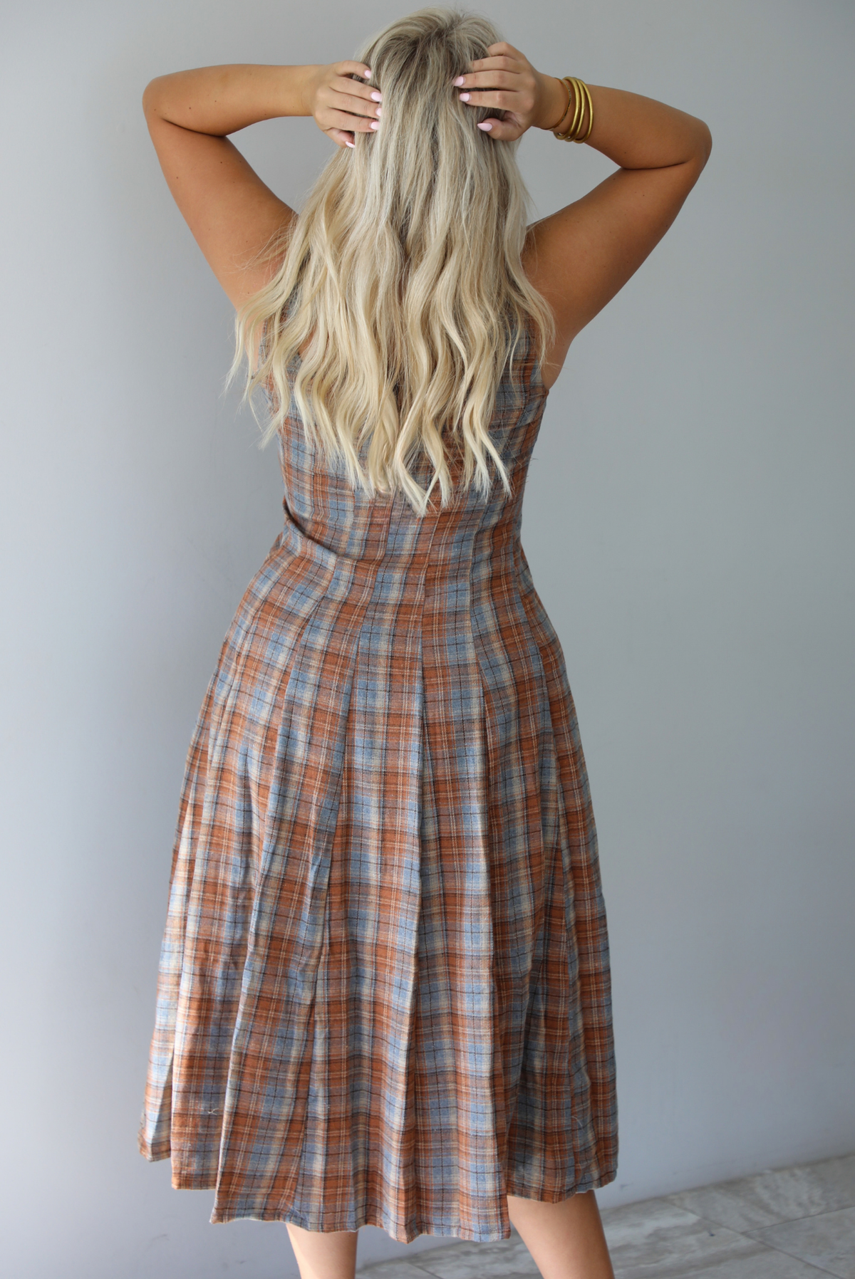 A woman shown from behind wearing a brown and blue plaid midi dress with thick straps, a square neckline, fitted bodice that starts flaring out at the hips and ends about mid-shin, with a flowy skirt, standing indoors against a white wall with white marble flooring.  