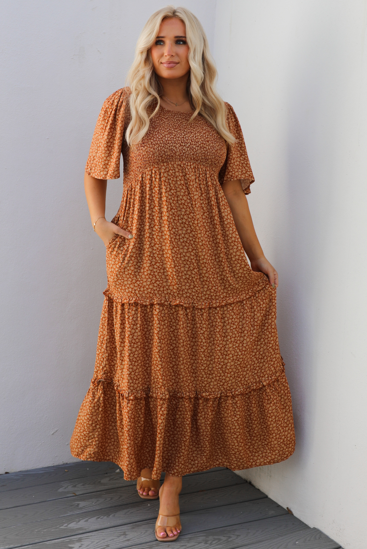 A woman model outdoors wearing a rust dress with a cream floral print pattern throughout. It has flowy short sleeves, a smocked bodice, and a flowy maxi length skirt with functional pockets on either side. 