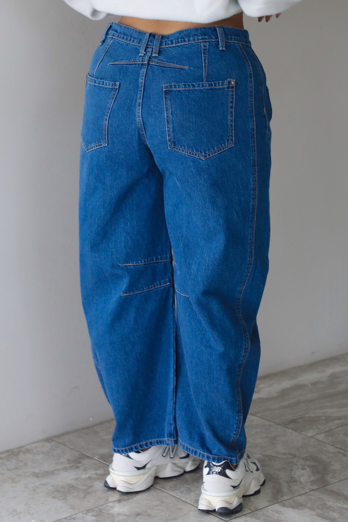 A woman shown from the waist down from behind, focusing on dark wash barrel denim jeans that have a mid-rise waistline, paired with a grey sweatshirt and New Balance chunky sneakers, standing indoors against a white wall with white marble flooring.  