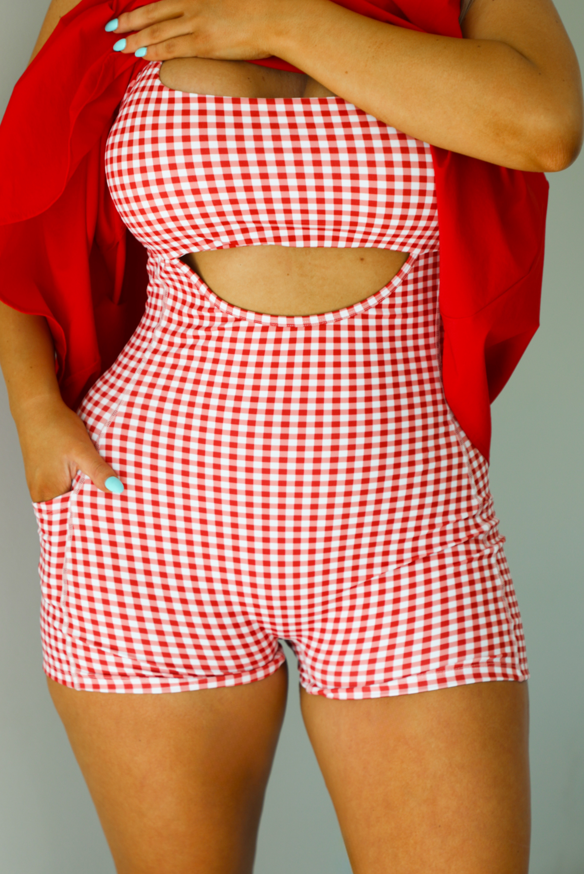 Woman wearing a red dress with a red and white gingham romper underneath. The photo is taken indoors against a white wall and shows a closeup of the romper underneath the dress, which has a cut out under the bodice and functional pockets on either side. 