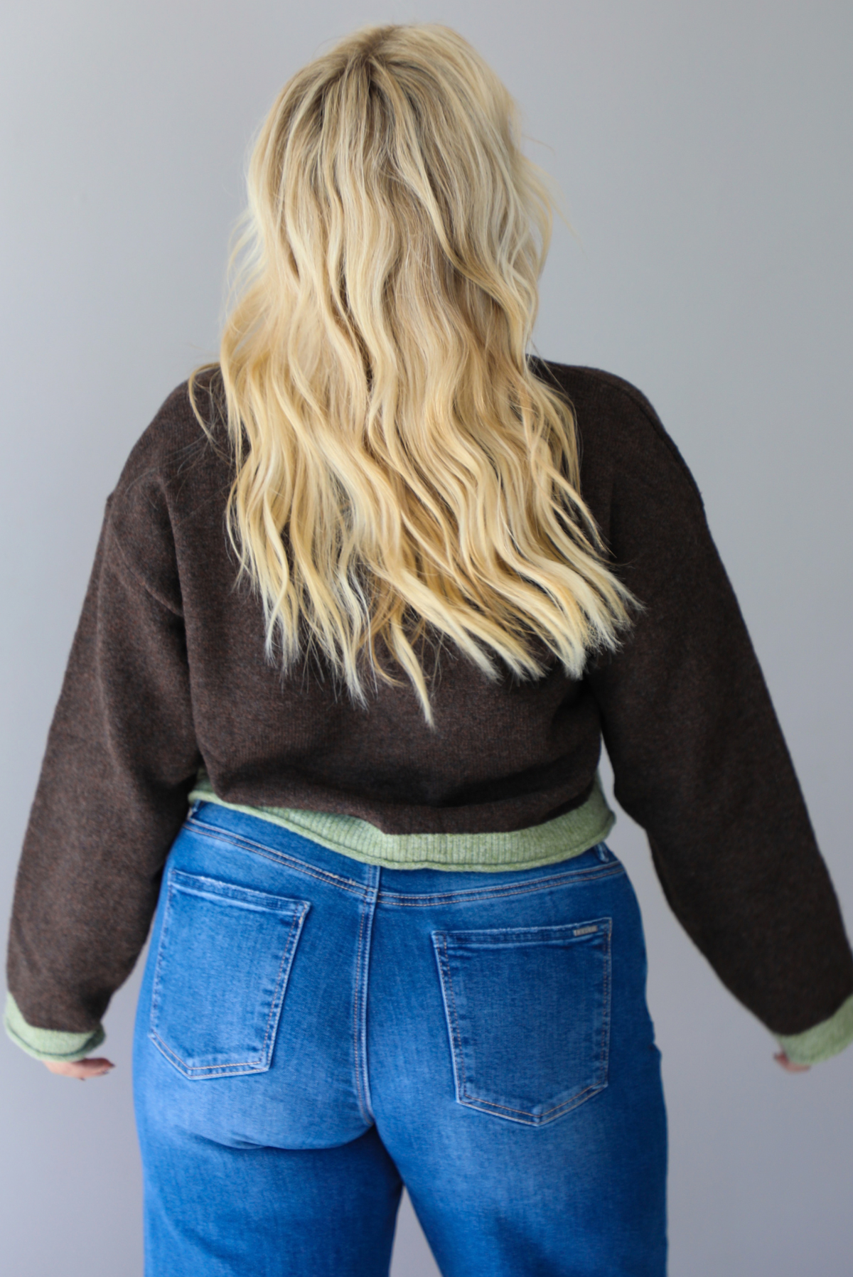 A woman shown from behind wearing a dark brown with green trim cardigan fully buttoned up paired with dark wash denim jeans, standing indoors against a white wall.