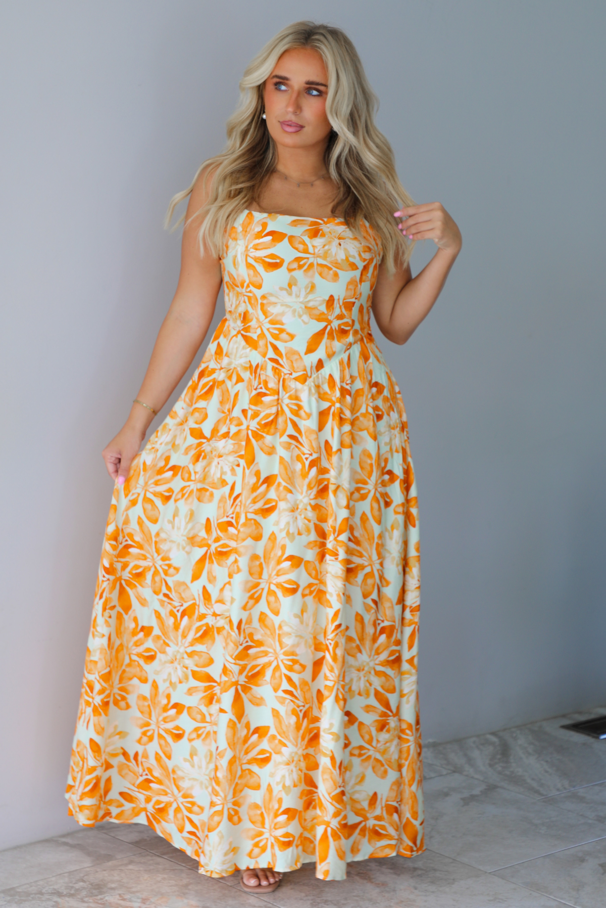 A woman modeling a spaghetti strapped, floor-length maxi dress with a white base and a vibrant marigold orange floral pattern, standing indoors against a white wall with white marble flooring. 